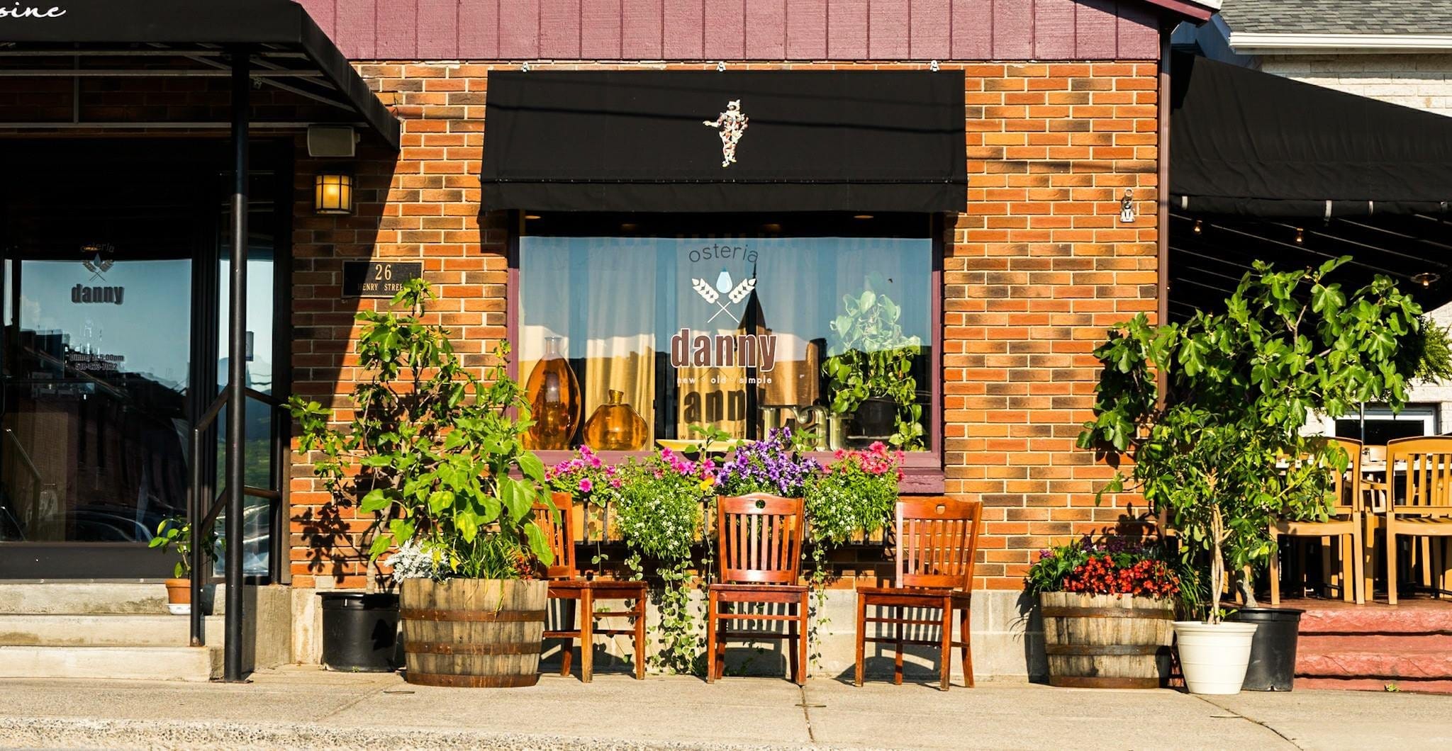 A cozy restaurant exterior with brick walls, large windows, and a black awning awaits near Saratoga Casino. Wooden chairs, potted plants, and colorful flowers decorate the inviting sidewalk in front.