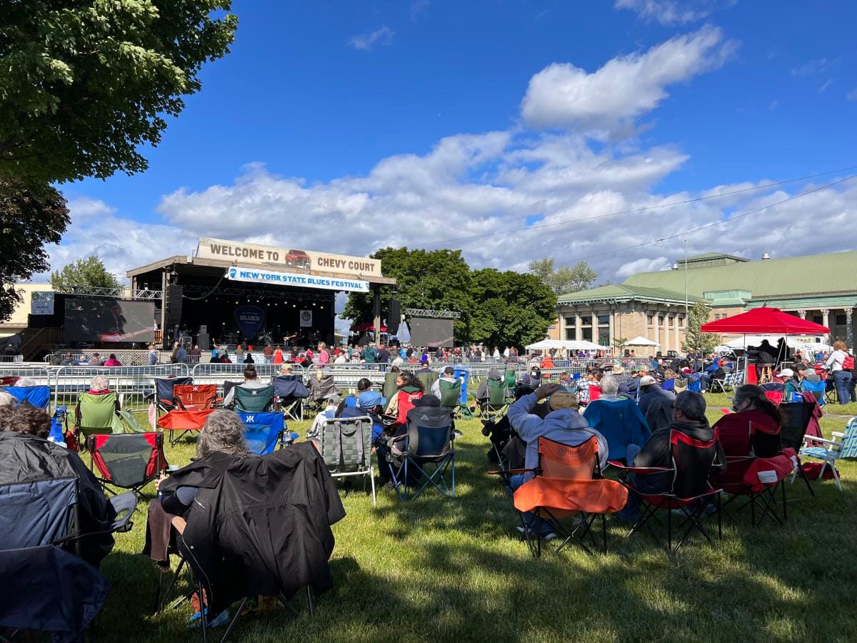 People sit on lawn chairs facing an outdoor stage at the New York State Fair’s Blues Festival, under a blue sky with scattered clouds. The stage has a sign reading “Chevy Court,” with trees and fair buildings in the background.