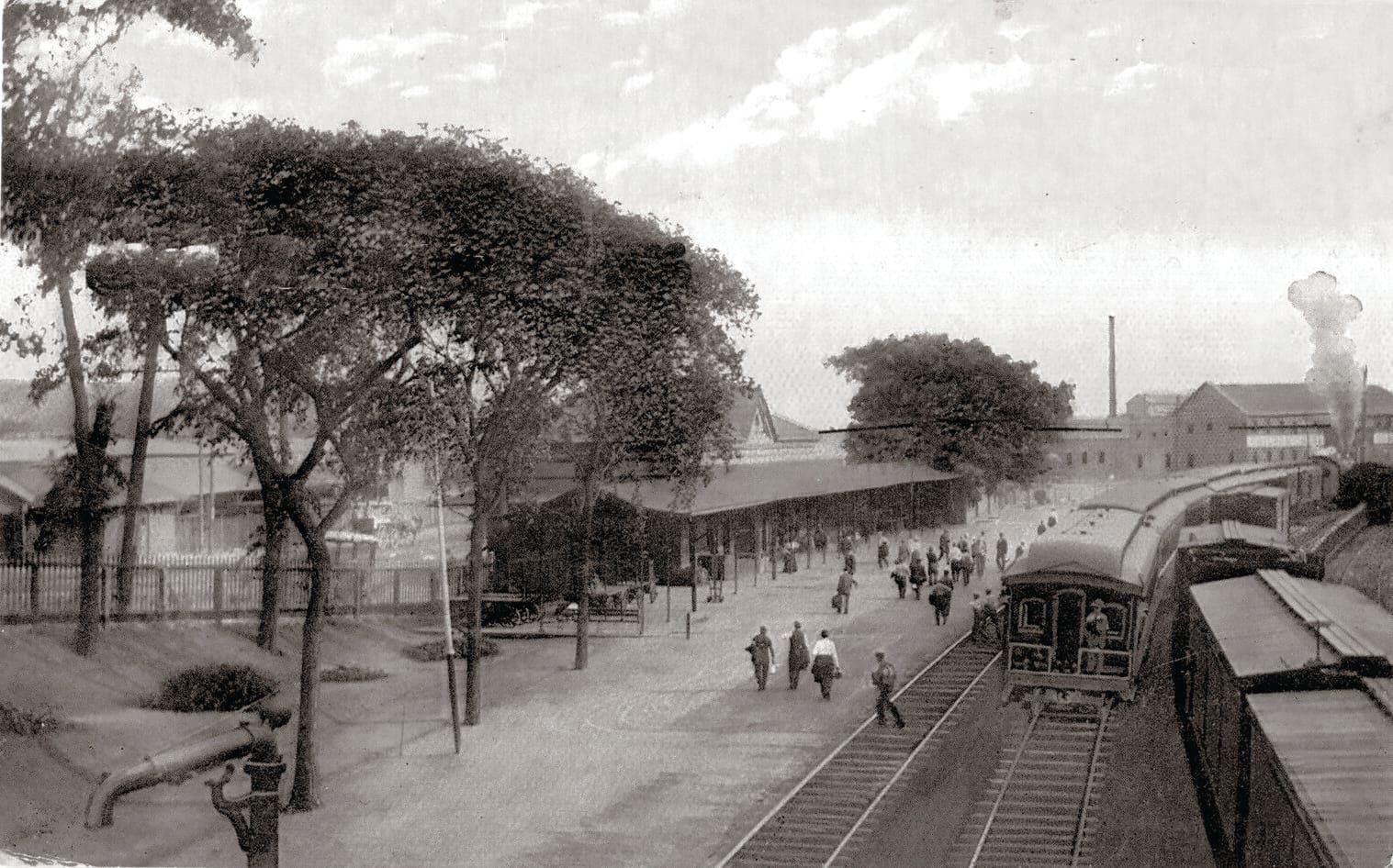Black-and-white photo of a train station platform with people walking and an Amtrak train on the tracks. Trees line the platform, and industrial buildings are visible in the background under a cloudy sky, hinting at the areas historic roots.