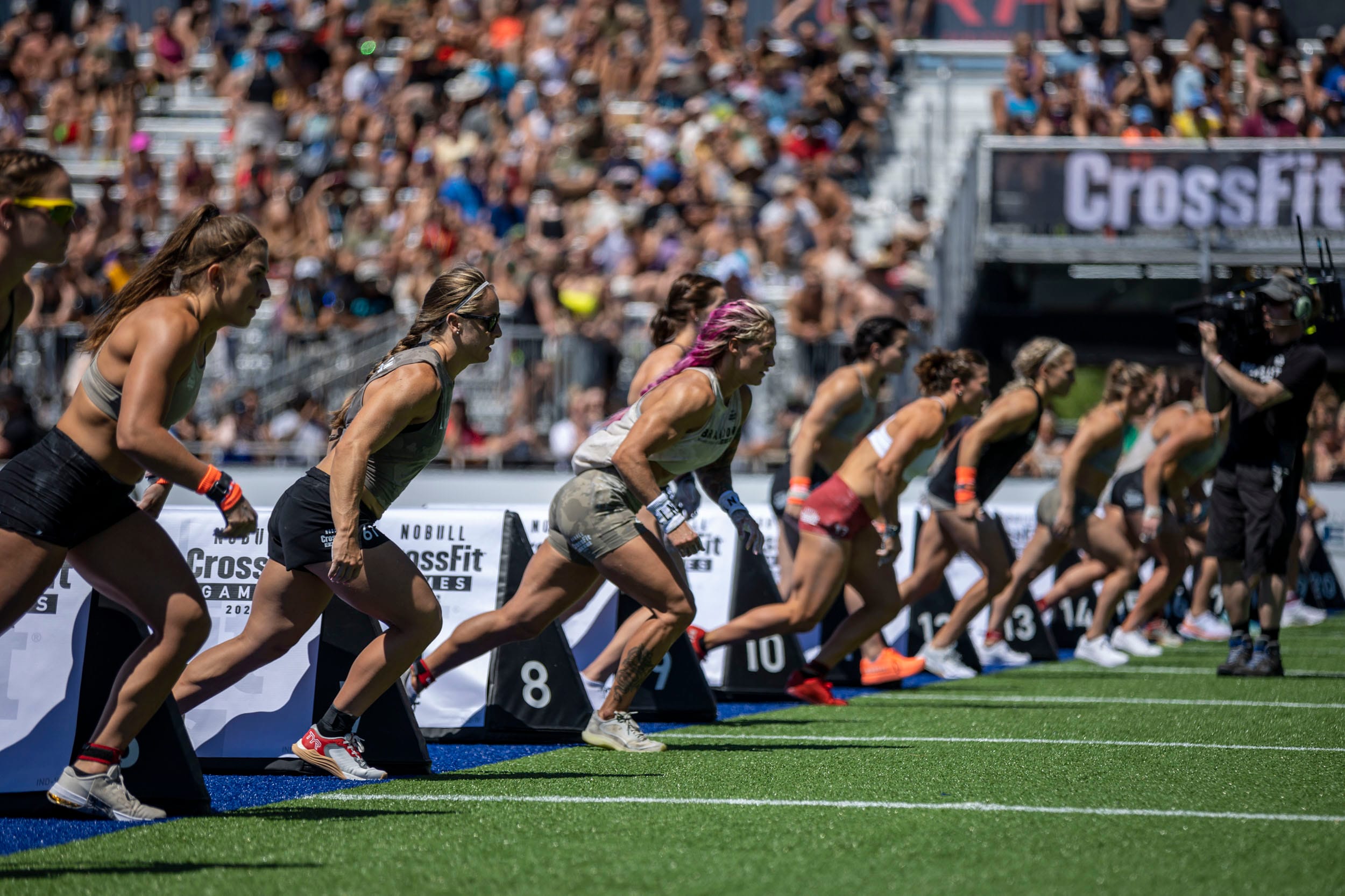 Several female athletes are lined up and starting a race on a turf field at a CrossFit competition, with a crowd of spectators and a large CrossFit sign—bringing the same energy youd find at a Flower & Garden Expo.