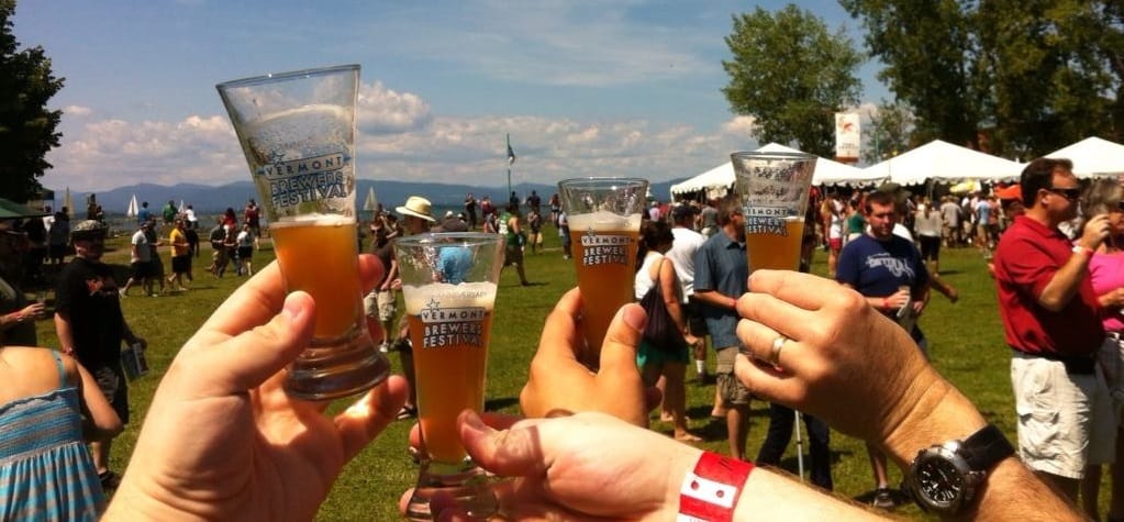 A group of people raise their beer glasses for a toast at the Vermont Brewers Festival, with tents, crowds, and trees visible in the background on a sunny day.