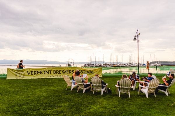 People sit in white chairs on a grassy lawn near a lake with sailboats, enjoying the Vermont Brewers Festival beneath an overcast sky. A lamppost stands nearby beside a large festival banner.