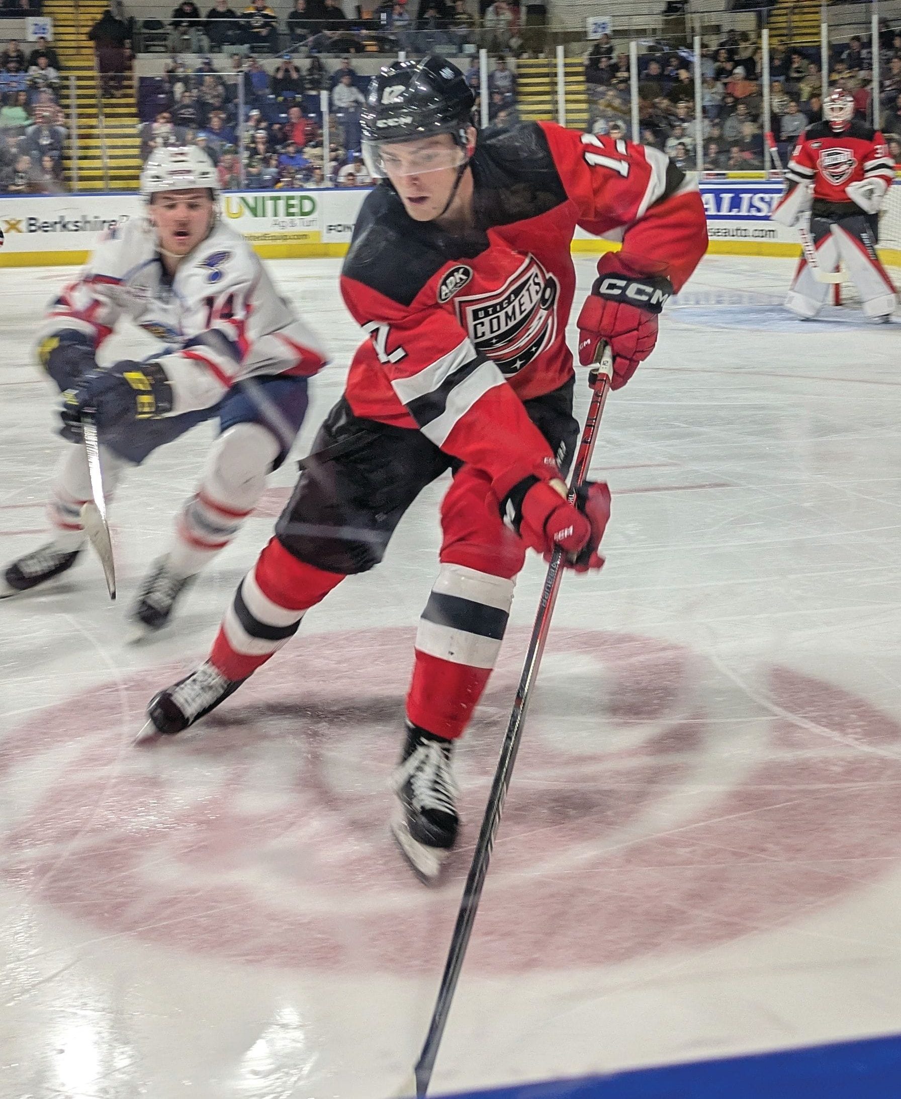 A hockey player in a red and black uniform controls the puck on the ice as a player in white skates behind him. The sports game is in full swing in a crowded indoor arena, capturing the excitement of live travel and competition.