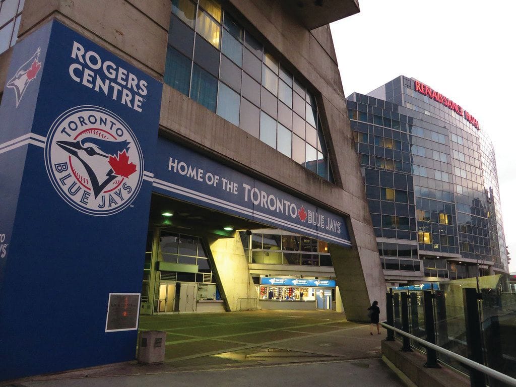 Entrance of Rogers Centre in Toronto, featuring Toronto Blue Jays signage and logo. A modern glass building labeled Renaissance Hotel is visible in the background—a quiet moment before a sports game draws crowds of fans.