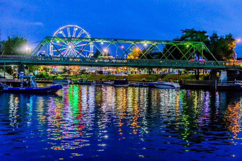 A brightly lit Ferris wheel and colorful lights from the Ticonderoga StreetFest reflect on the water beneath a green metal bridge at night, with boats docked along the river and trees in the background.