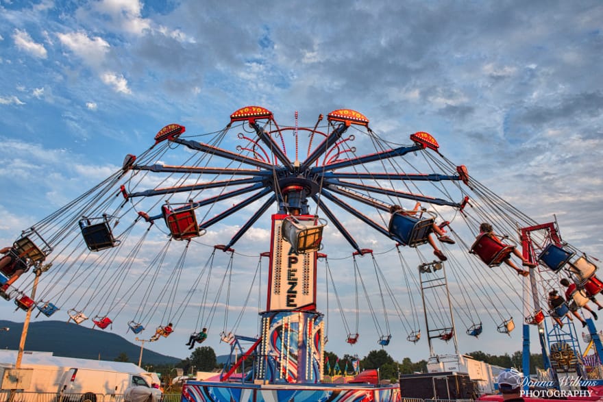 A colorful swing ride at a fair spins with empty and occupied seats under a blue sky, as mountains and carnival booths—including those from the Vermont Brewers Festival—dot the scenic background.