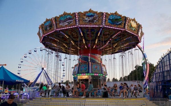 People enjoy a colorful swing carousel at the Dutchess County Fair during dusk, with a Ferris wheel and game booths in the background, and trees silhouetted against the evening sky.