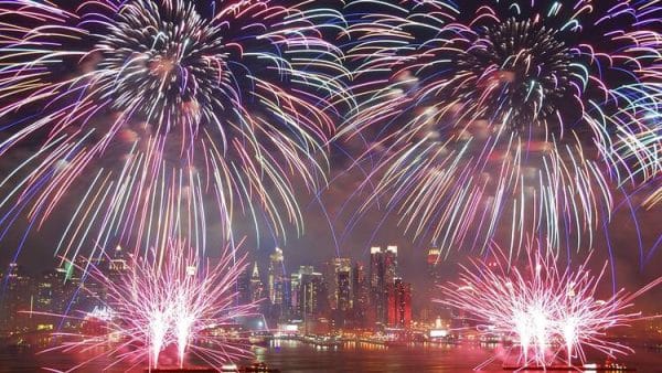 Colorful 4th of July Fireworks burst over a city skyline at night, illuminating the buildings and reflecting on the water below. The sky is filled with vibrant streaks of light in pink, purple, and blue.