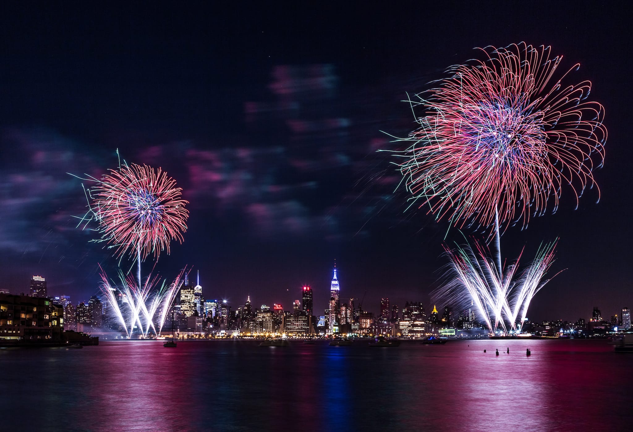 4th of July Fireworks in red, white, and blue burst over a city skyline at night, reflecting in the water below. The illuminated buildings add to the festive and vibrant atmosphere.