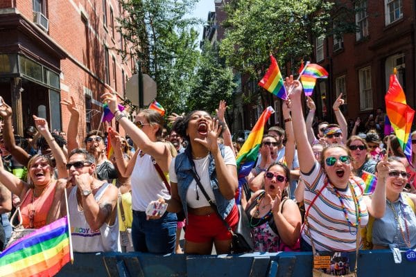 A diverse, energetic crowd waves rainbow flags and cheers behind a blue barrier at an outdoor Pride parade on a sunny day, celebrating LGBTQ+ pride and unity on a city street lined with brick buildings and trees.