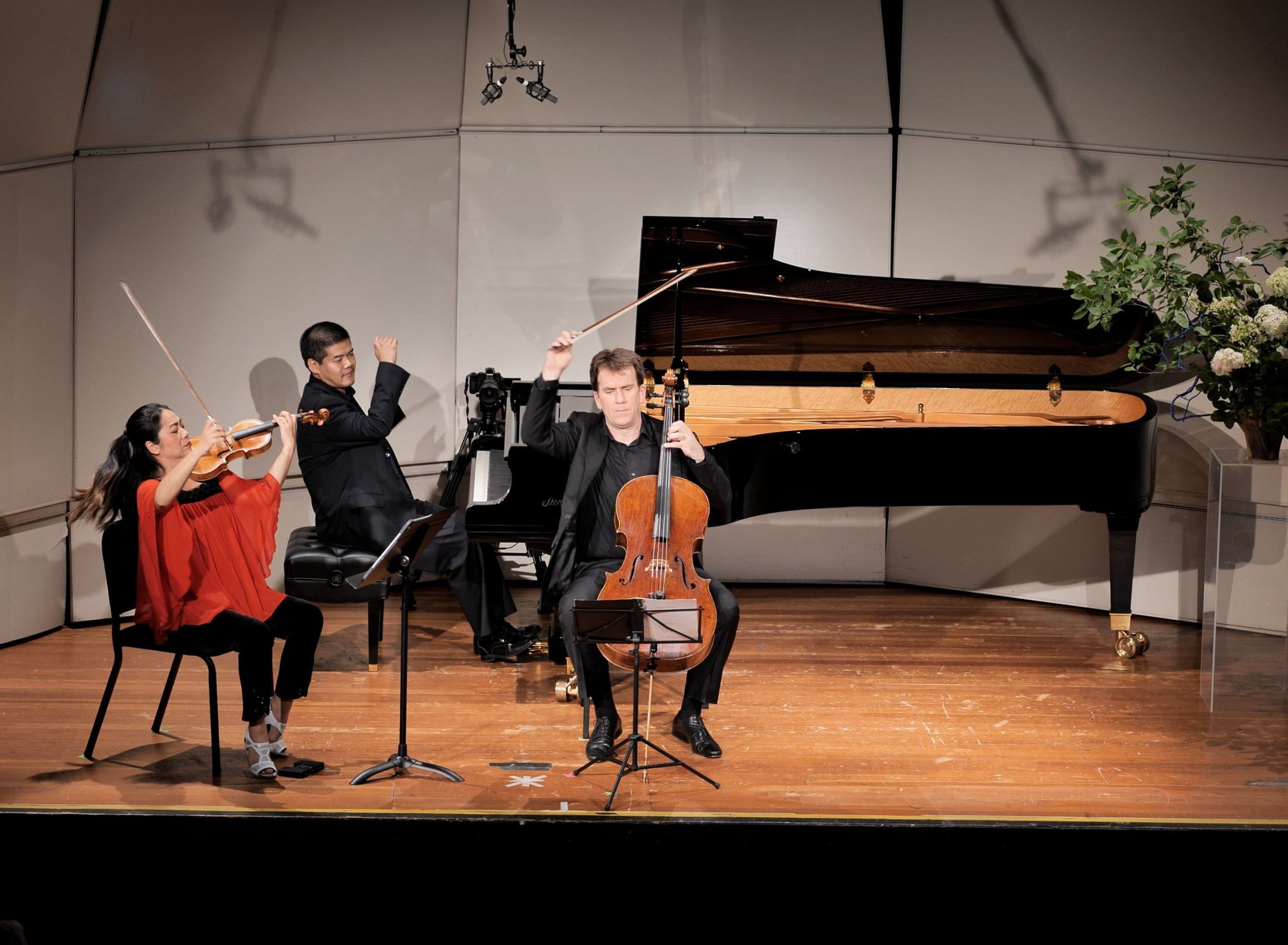 Three musicians perform on stage at the Manchester Music Festival: a woman playing violin, a man at a grand piano, and another man playing cello, all mid-performance with music stands and sheet music in front of them.