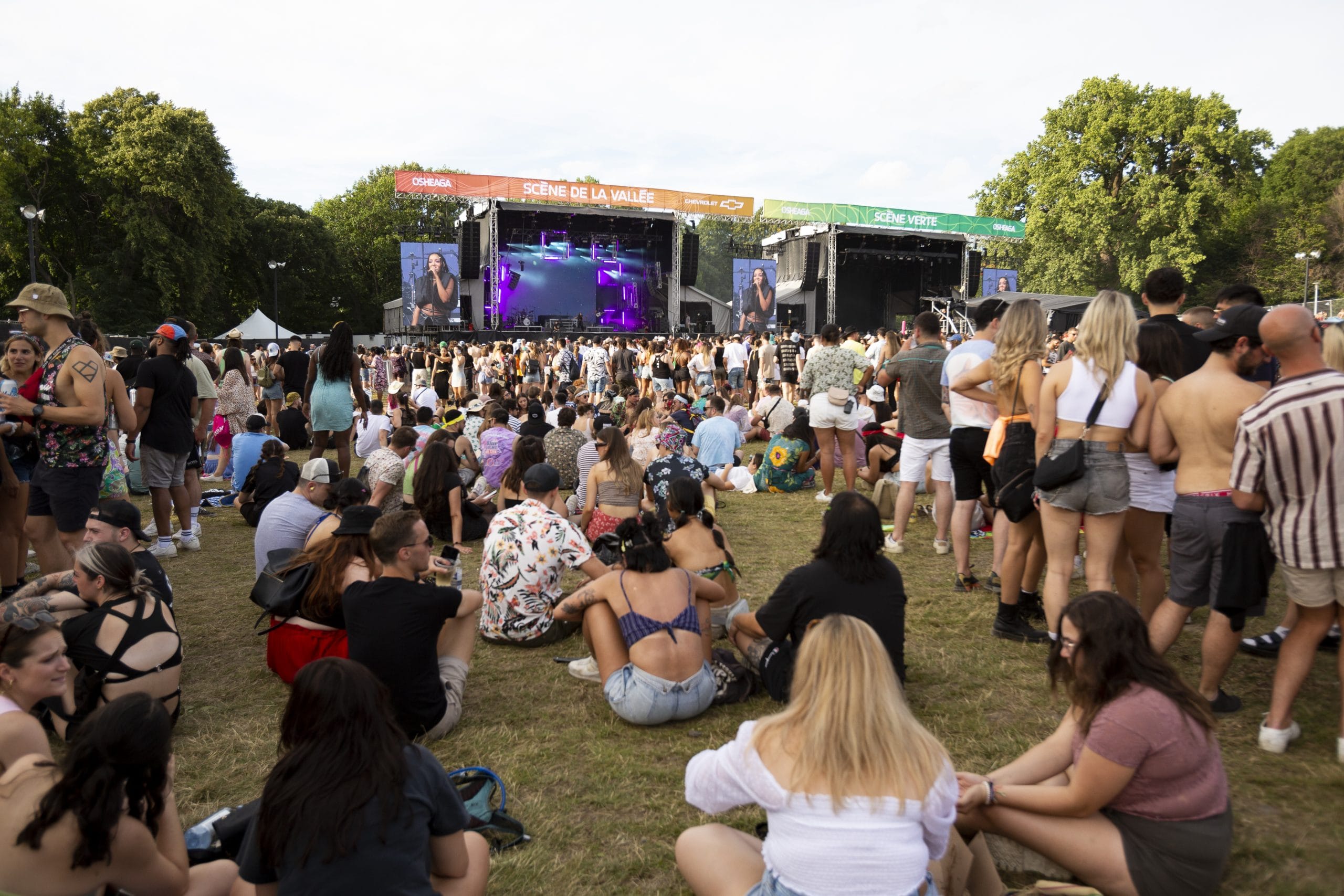 A large crowd of people sits and stands on grass at the Osheaga outdoor music festival, facing a stage with colorful lights and performers. Trees surround the area, and it is daytime with clear weather.