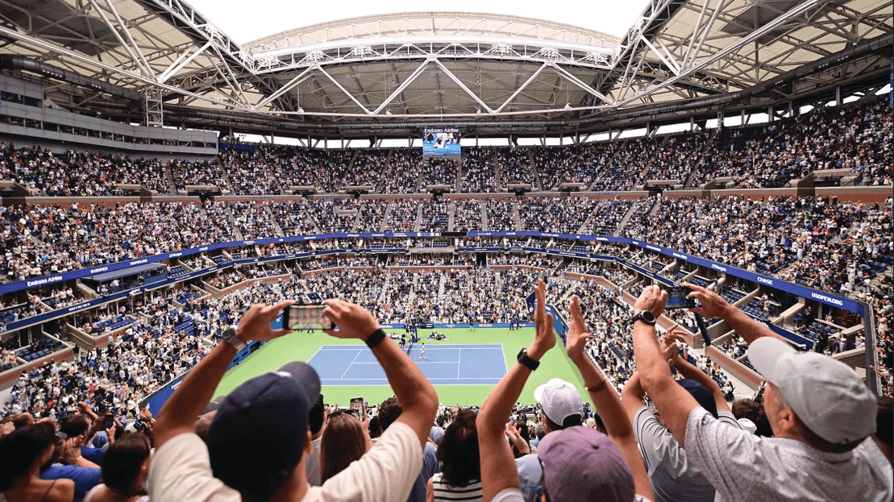 A large crowd travels to the stadium, cheering and taking photos while watching a thrilling sports game on a blue tennis court under a partially open roof. The stands are filled with excited spectators, many raising their hands in excitement.