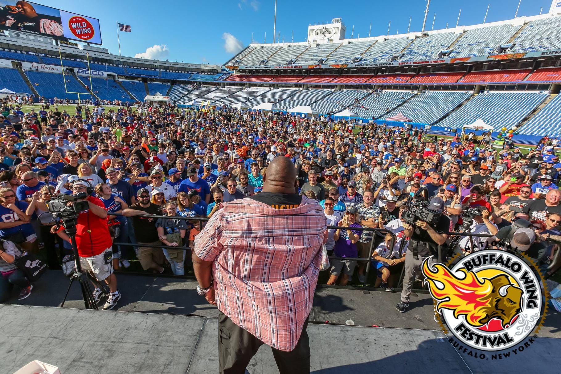 A man addresses a large crowd at the Buffalo Wing Festival in Buffalo, New York. People in casual attire gather near the stage, surrounded by stadium seats and blue sky in the background.