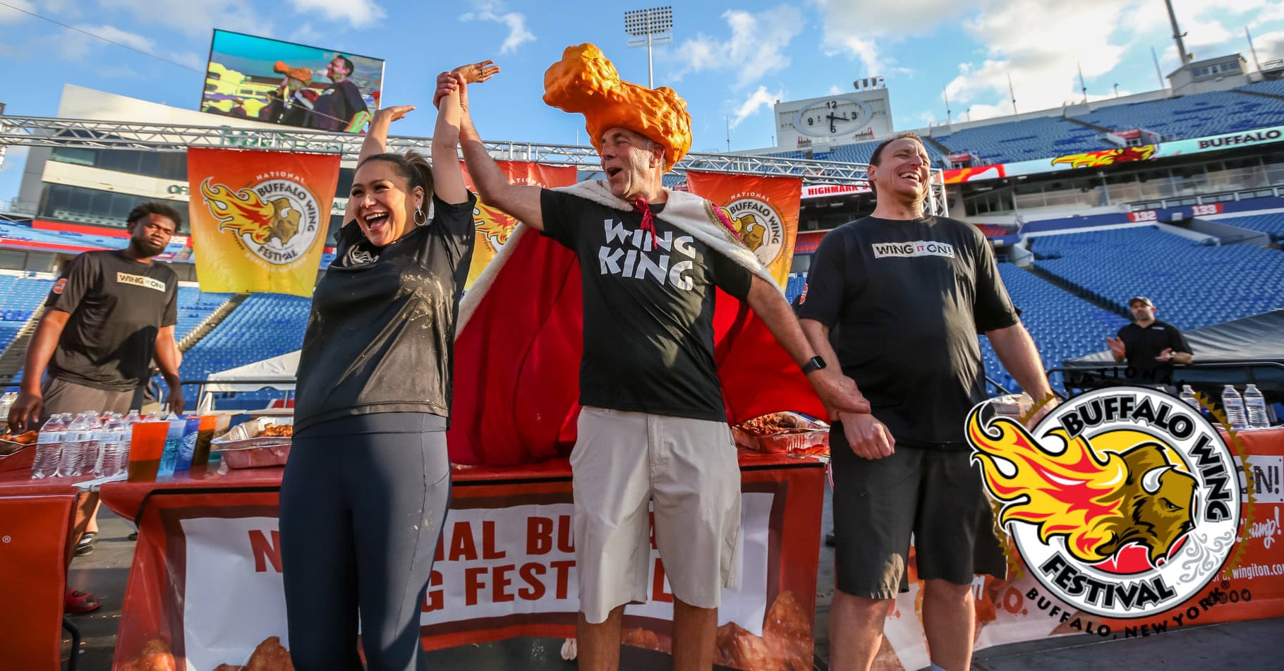 Three people celebrate on stage at a stadium during the Buffalo Wing Festival, one sporting a large chicken wing hat and a Wing King shirt. The Buffalo Wing Festival logo stands out in the foreground, with banners and tables of food nearby.