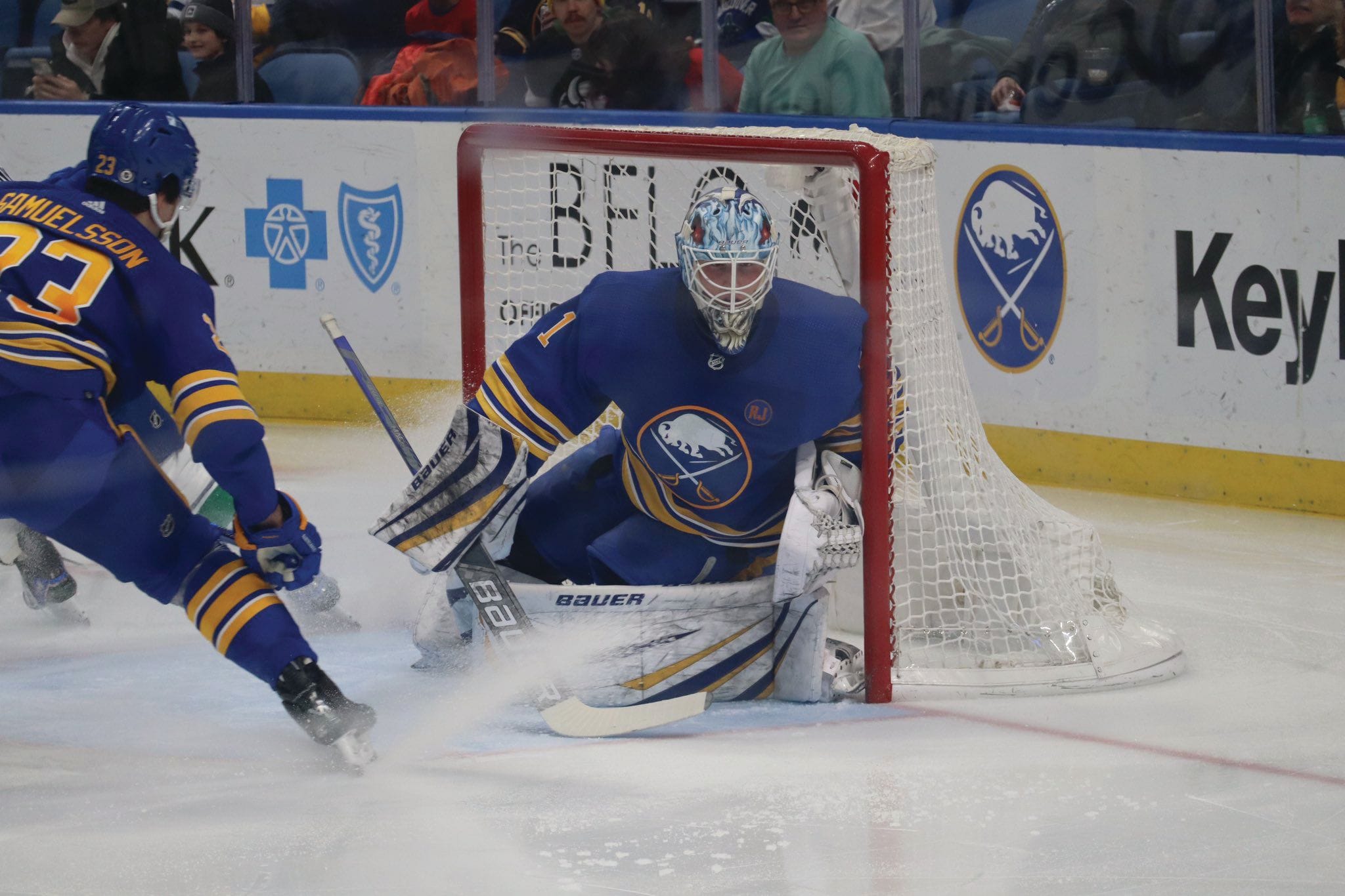 A hockey goalie in blue and yellow gear guards the net as a teammate skates nearby during a sports game. Spectators watch from the stands behind the glass, perhaps having taken Amtrak to enjoy the action.