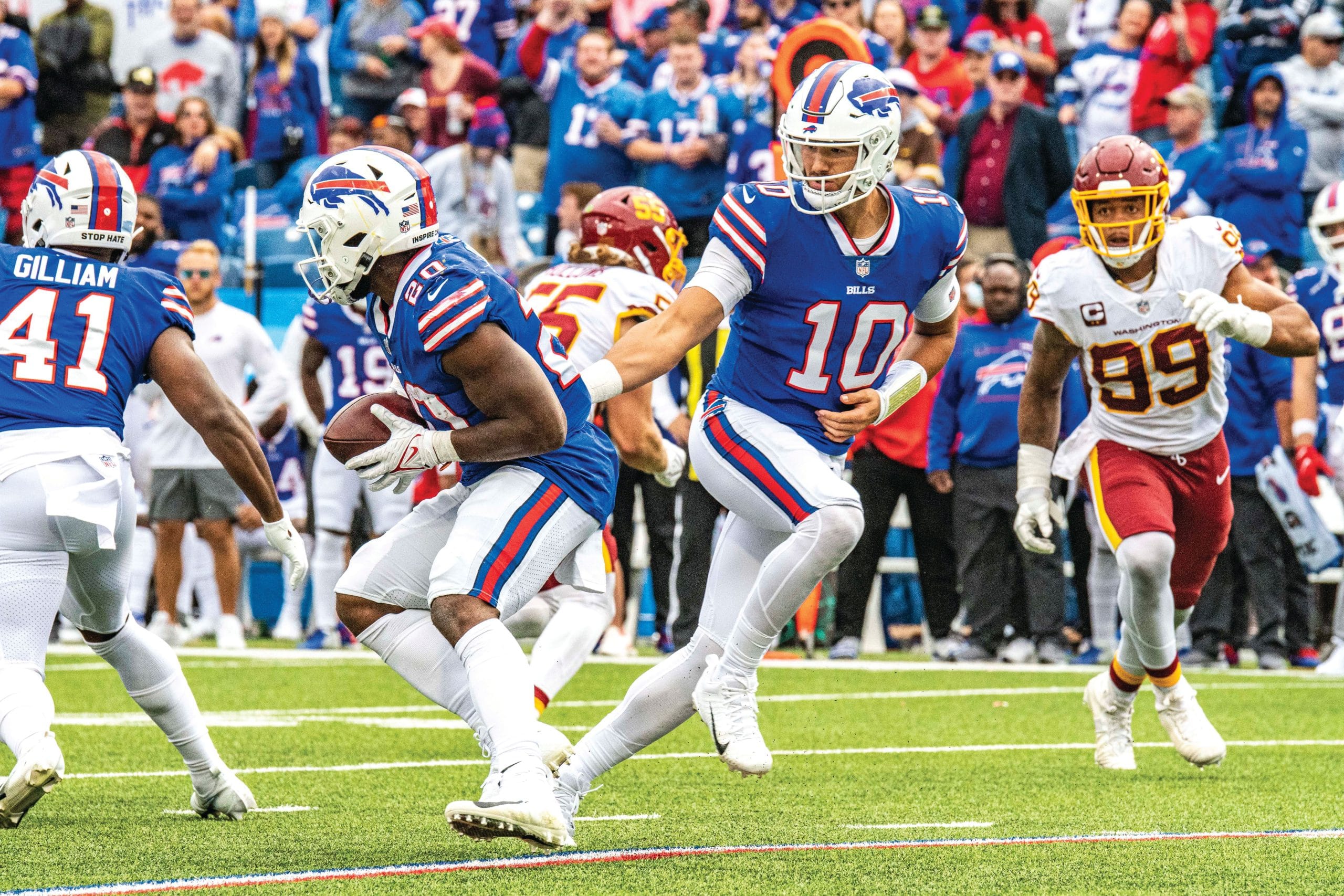 During a sports game, a football quarterback in a blue jersey hands the ball to a running back as opposing players in white and red approach, with a crowd of fans in the background—capturing the excitement of travel and competition.