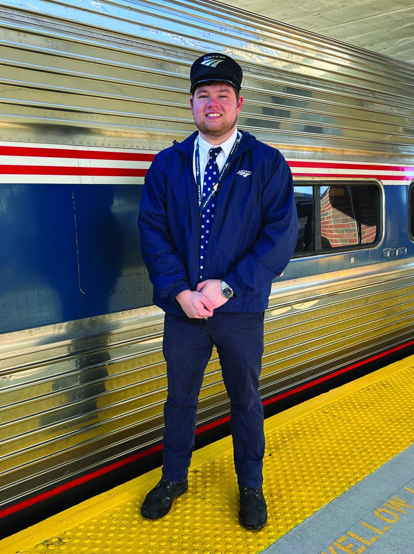 A smiling train conductor in uniform stands on a platform next to a silver Amtrak train, showing pride in his conductor career. The yellow safety line marked YELLOW LINE runs along the ground beside him.