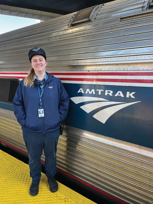 A smiling Amtrak conductor in uniform and cap stands on a yellow platform next to an Amtrak train, proudly representing the conductor career with the silver exterior, red, white, and blue stripes, and iconic Amtrak logo.