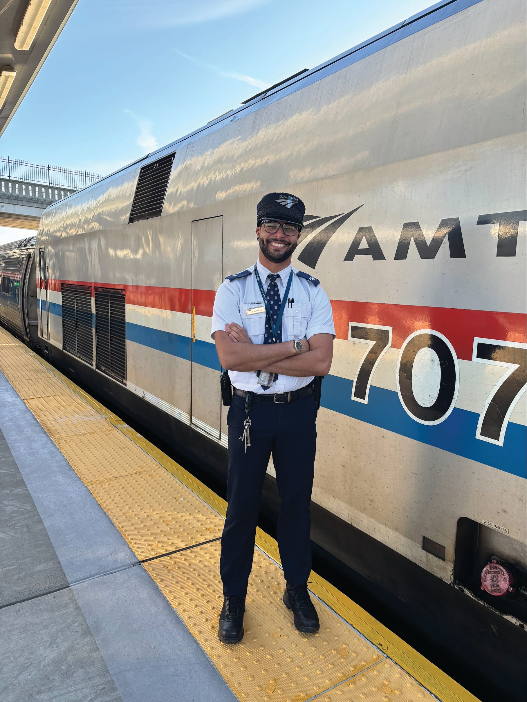 A smiling Amtrak conductor in uniform stands with arms crossed on a yellow platform next to an Amtrak train at a station, embodying the confidence and leadership seen in conductors pursuing a music career, with blue sky in the background.
