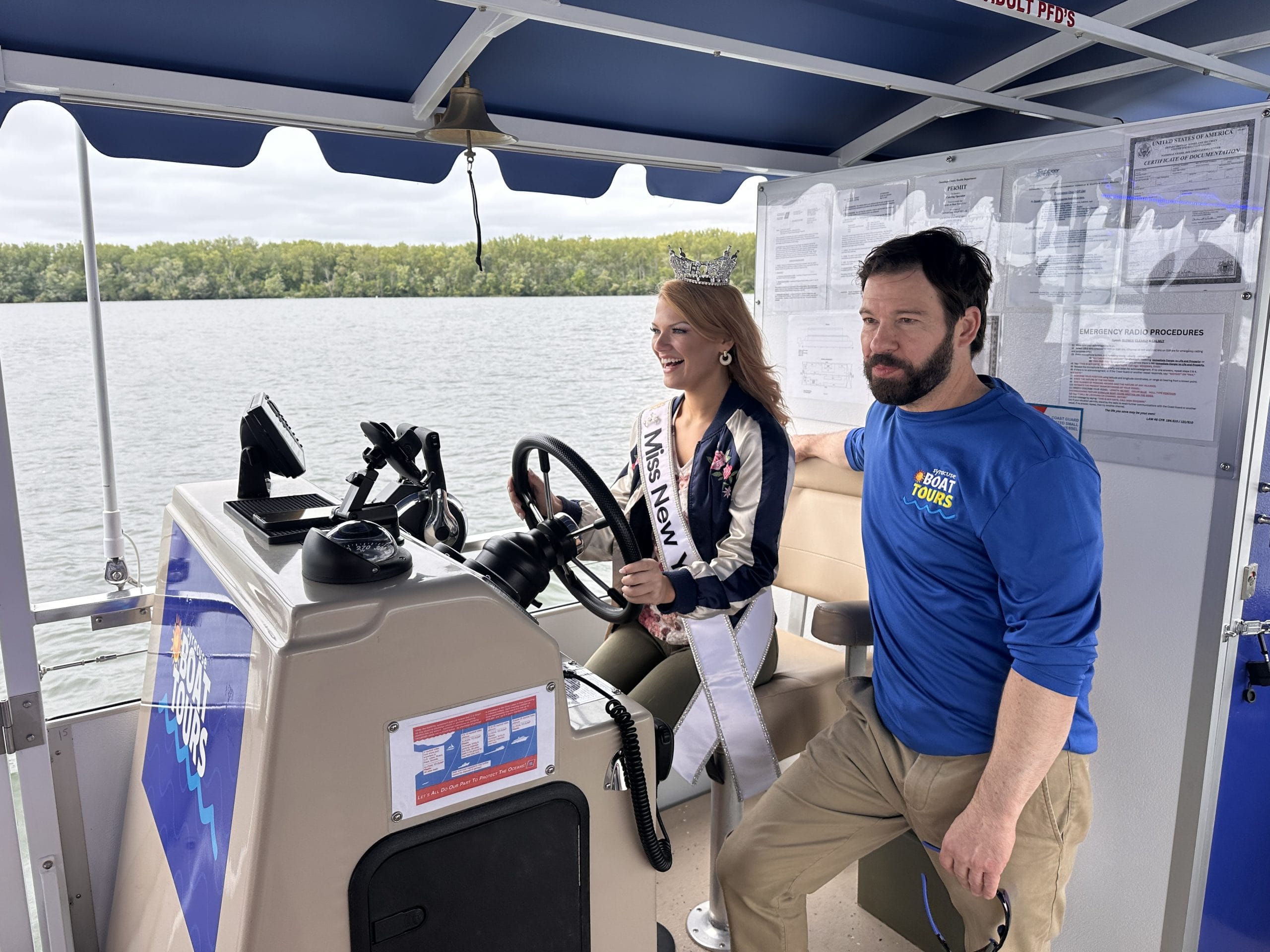 A woman wearing a crown and sash, crowned Syracuse Miss New York, sits at the wheel of a boat, smiling, while a man in a blue shirt stands beside her. They are on a lake with trees visible in the background.