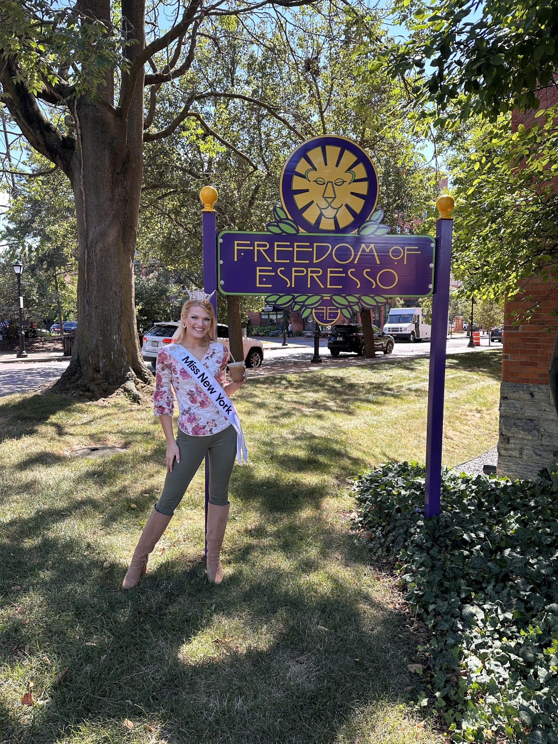 A woman, crowned Syracuse Miss New York, stands proudly in front of a sign.