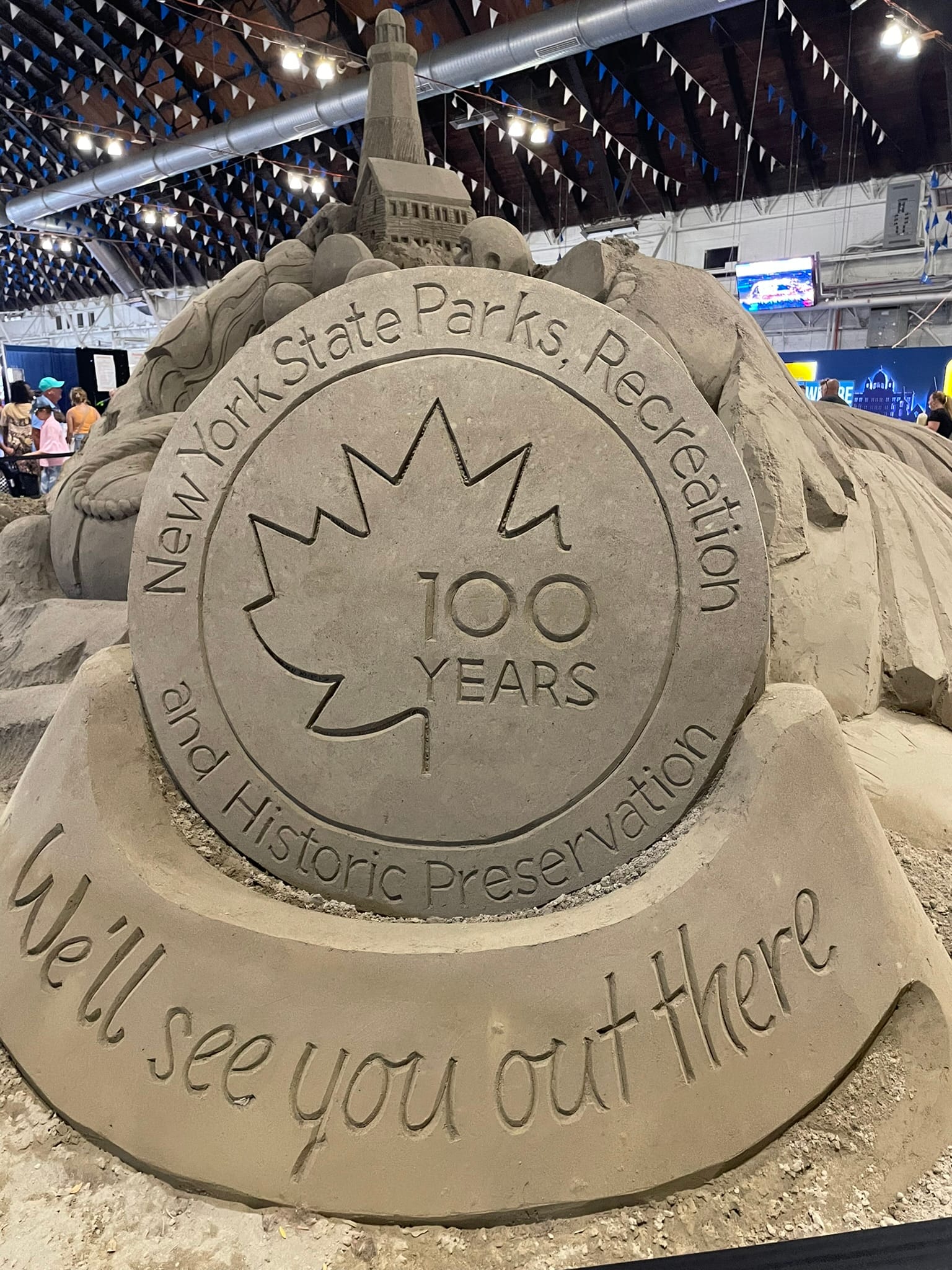 A detailed sand sculpture displays a large medallion with a maple leaf and text reading New York State Parks, Recreation and Historic Preservation 100 Years. Below, it says, We’ll see you out there—explore even without a car.