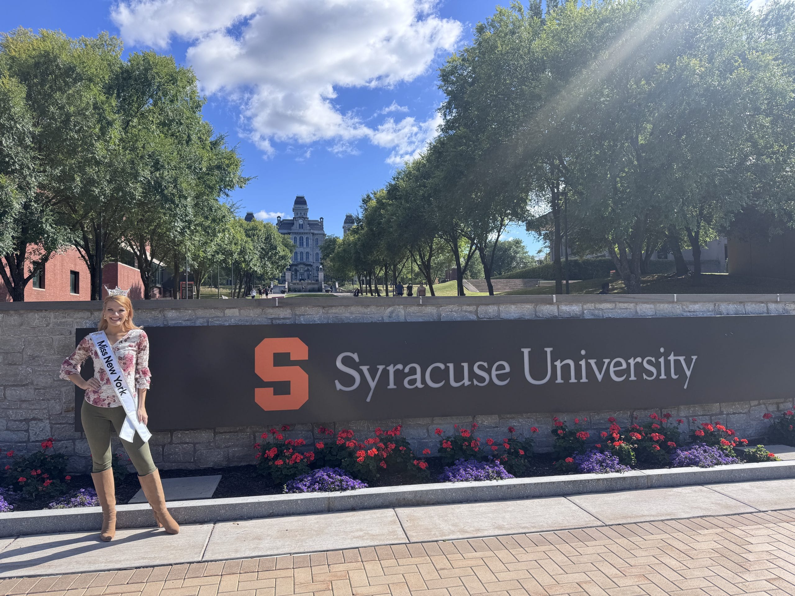 A woman wearing a sash and crown stands and smiles in front of a Syracuse University sign, surrounded by flowers. Representing Miss New York, she poses with trees and campus buildings behind her under a partly cloudy sky.