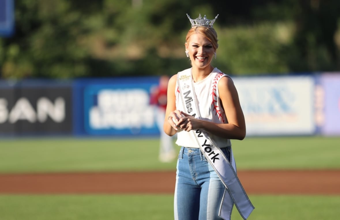 A woman wearing a crown, sash reading Miss New York, white top, and jeans smiles while standing on a Syracuse Mets baseball field. The background is blurry with a green outfield and advertising signs.