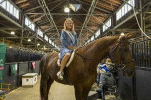 A smiling woman, crowned Syracuse Miss New York, wearing a sash and dress, sits on a brown horse inside a stable with wooden beams and metal railings. Other people and stable equipment are visible in the background.
