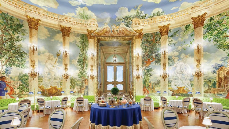 An ornate, light-filled dining room at the Pierre Hotel in NYC features high ceilings, marble columns, opulent chandeliers, and lush classical garden scenes. Elegant tables are arranged neatly, with a central table set for tea.