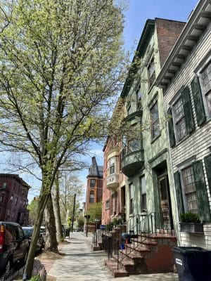A sunny street scene shows colorful historic row houses, with green, white, and red facades, beside a large leafy tree and parked cars. A taller brick building with a pointed roof is visible in the background.