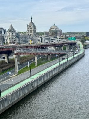 A riverside view of a city shows a multi-level road with a green bike and pedestrian path, light traffic, and distinctive historic buildings in the background under a cloudy sky.