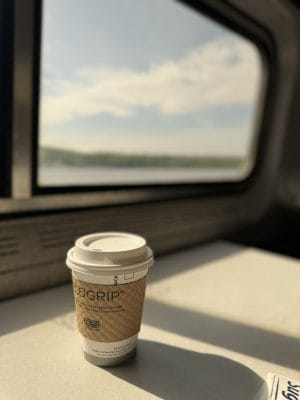 A disposable coffee cup with a brown sleeve sits on a table by a sunlit train window, with a blurred landscape and sky visible outside.