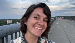 A woman with short brown hair and a striped shirt smiles while taking a selfie on a bridge without a car in sight. The sky is mostly cloudy, and traffic cones line the empty pedestrian path.