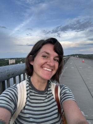 A person with short brown hair and a striped shirt smiles while taking a selfie on a wide, car-free pedestrian bridge under a partly cloudy sky. Orange cones line the pathway in the background, highlighting the space without a car in sight.