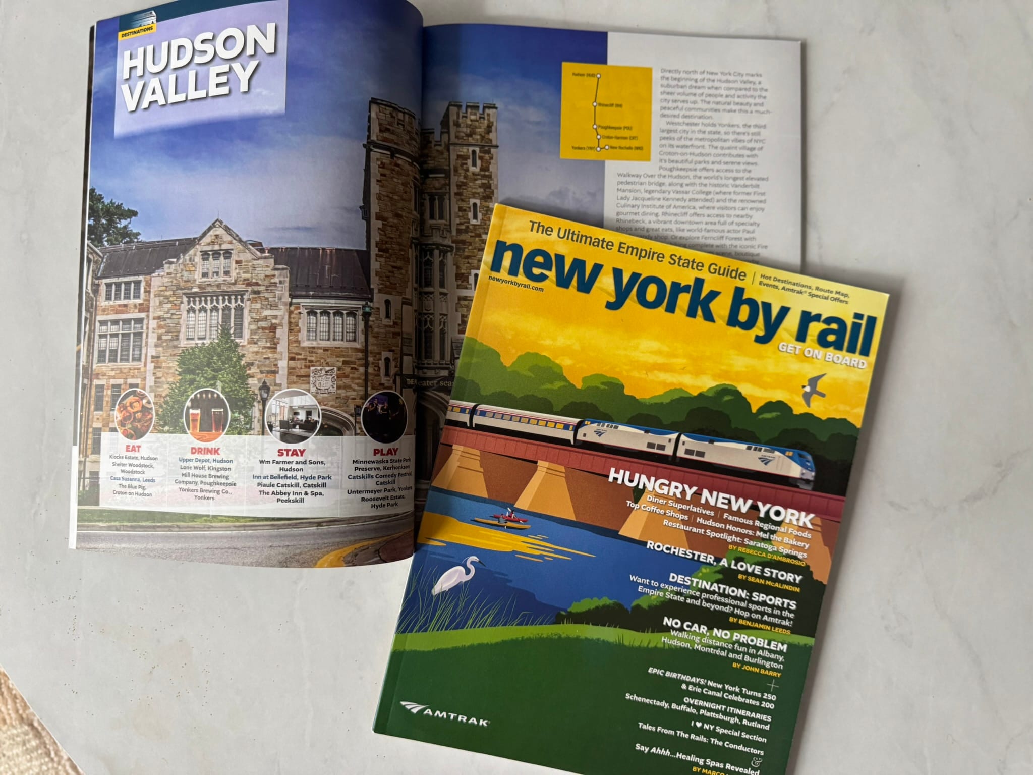A magazine titled new york by rail lies on a marble surface, partially covering an open page featuring Hudson Valley with a photo of a large stone building and sections labeled Eat, Drink, Stay, and Play—perfect for exploring without a car.