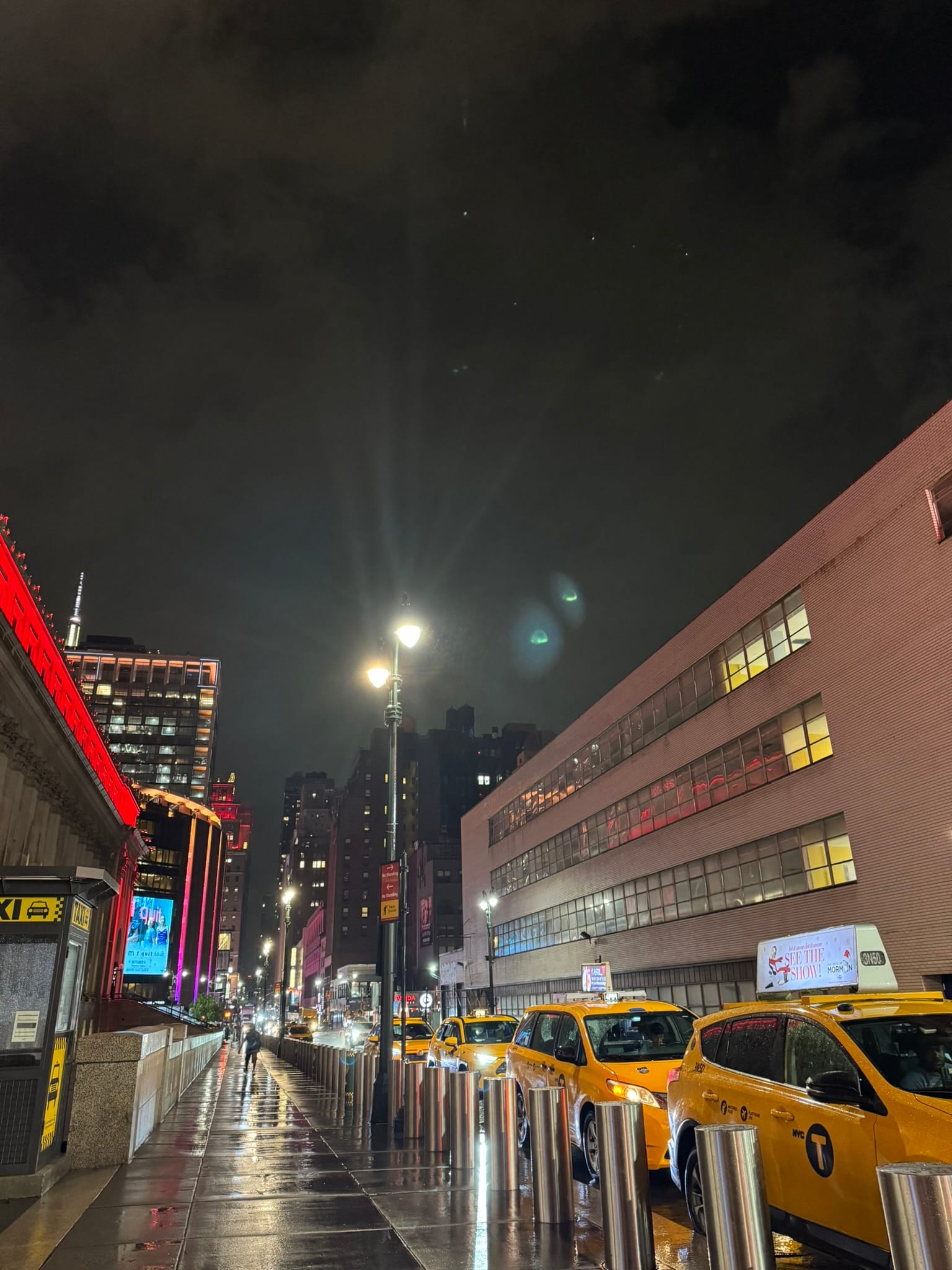 A rainy night in New York City unfolds without a car, with yellow taxis lined up along a wet street, streetlights reflecting on the pavement, and tall buildings illuminated in the background.
