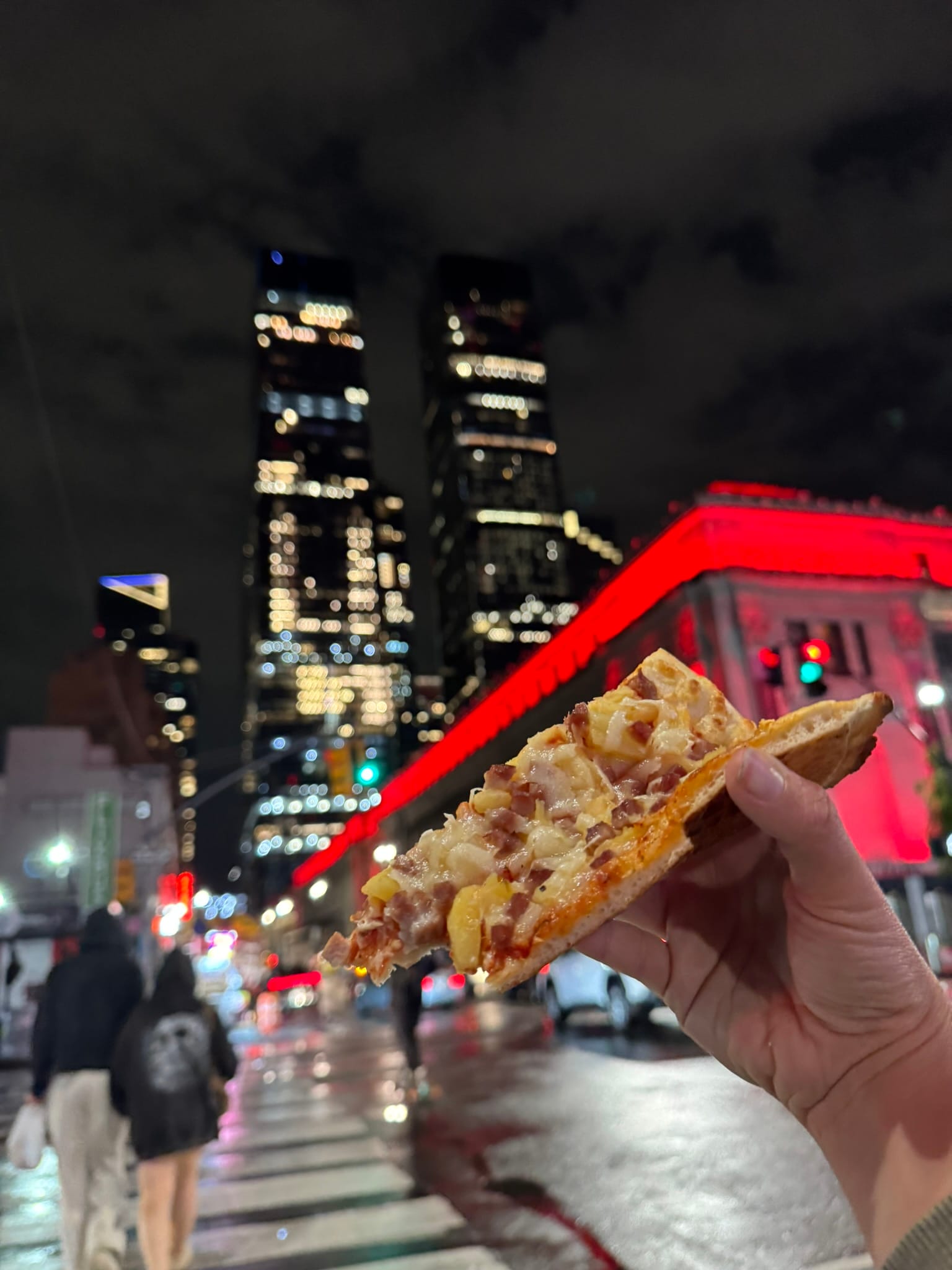 A hand holds a slice of pizza in the foreground, while people stroll without a car along a bustling city street lined with tall, illuminated skyscrapers. A building glows red beneath the night sky.