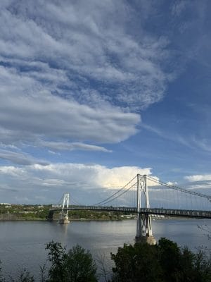 A suspension bridge stretches across a wide river under a partly cloudy blue sky, with green trees in the foreground and distant buildings along the riverbank—an inviting scene to explore without a car.