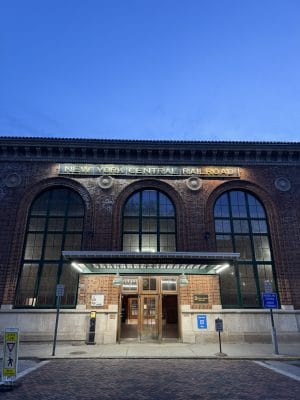 A red brick building with large arched windows displays a lit-up New York Central Rail Road sign above the entrance. The clear dusk sky sets the scene, and several signs near the entrance make it easy to find—even without a car.