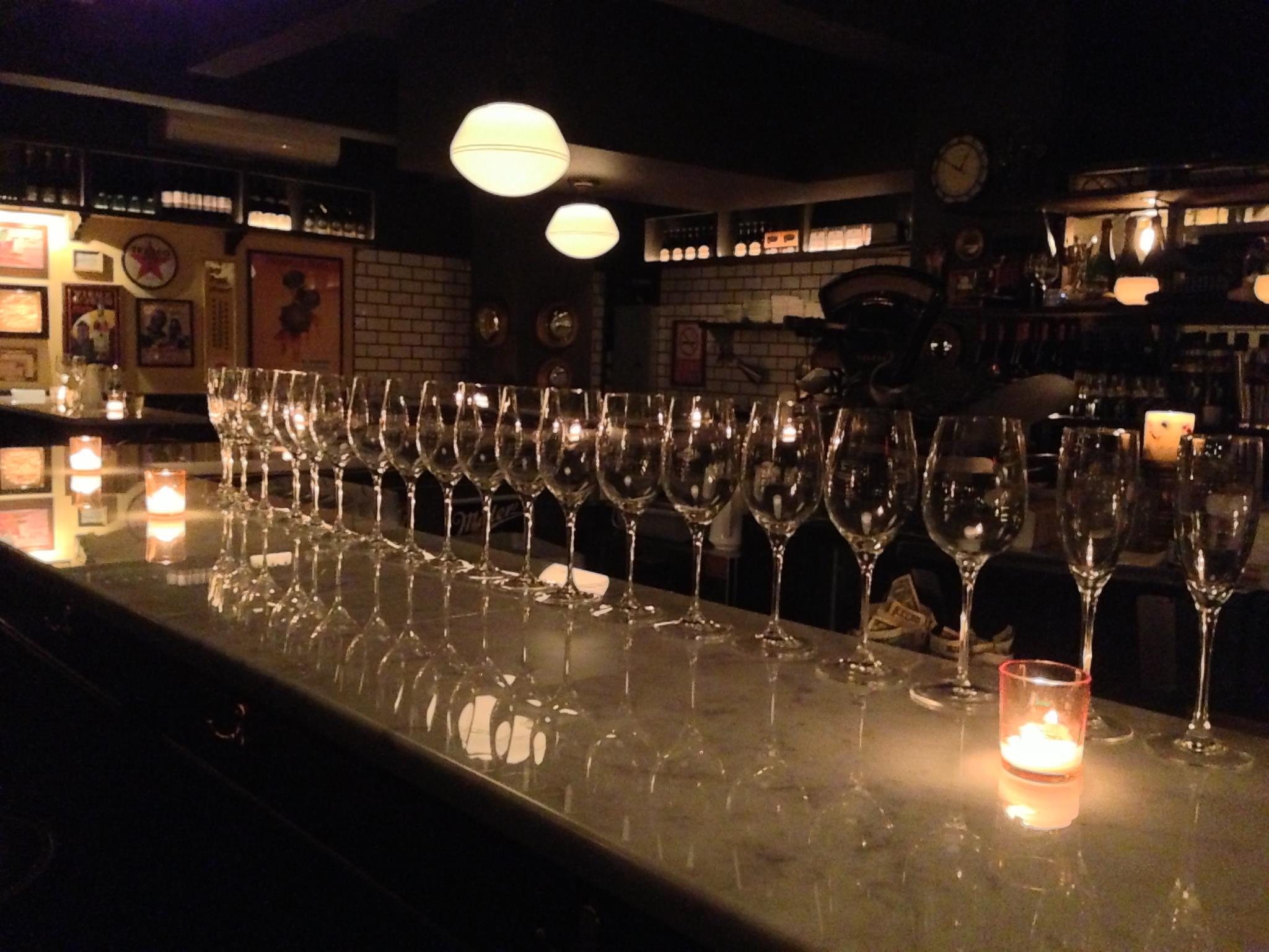 A dimly lit bar at the Vanguard Wine Bar features a row of empty champagne glasses on a polished counter, illuminated by small candles and soft overhead lights, with shelves and framed pictures in the background.