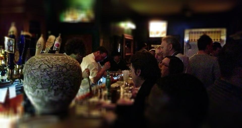 A dimly lit bar at 9 Maple Ave buzzes with a lively, social crowd gathered around the counter, as bartenders craft drinks and a large decorative vase stands prominently in the foreground.