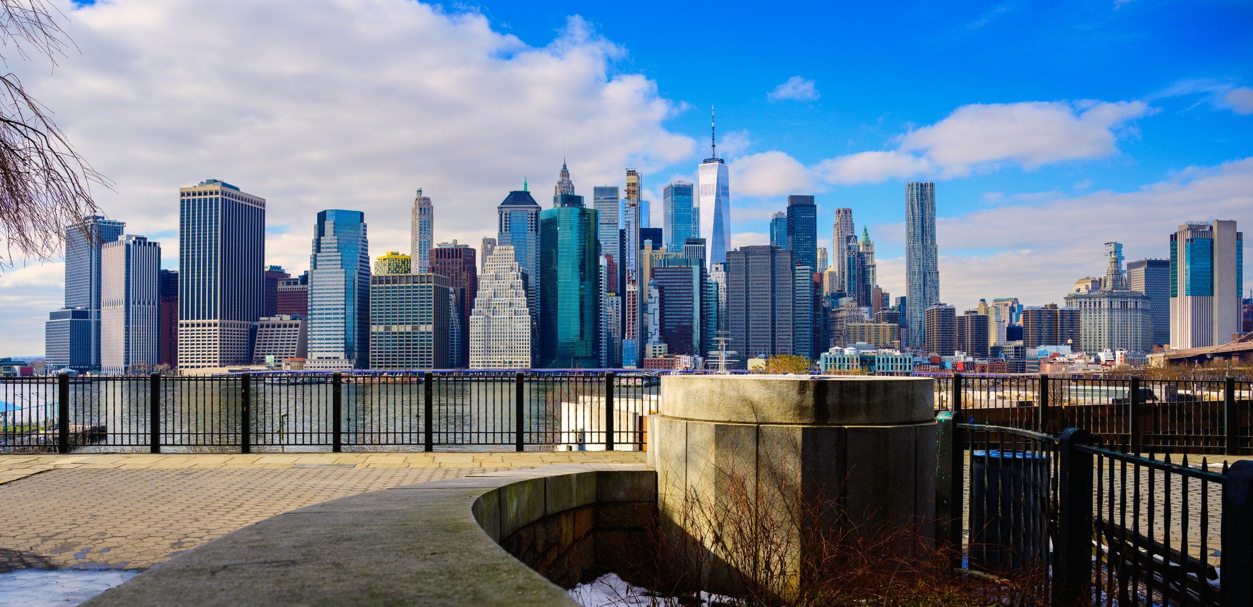 View of lower Manhattan skyscrapers from across the East River, with a blue sky, partial clouds, and the Brooklyn Heights Promenades railing and concrete structure in the foreground.