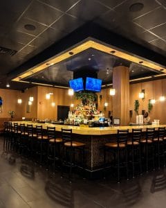 A modern bar with a square counter surrounded by high chairs, illuminated by hanging pendant lights. Bottles and TVs line the back, and a single person sits at the far end. The warm, inviting atmosphere makes Ember and Cork the perfect gathering spot.