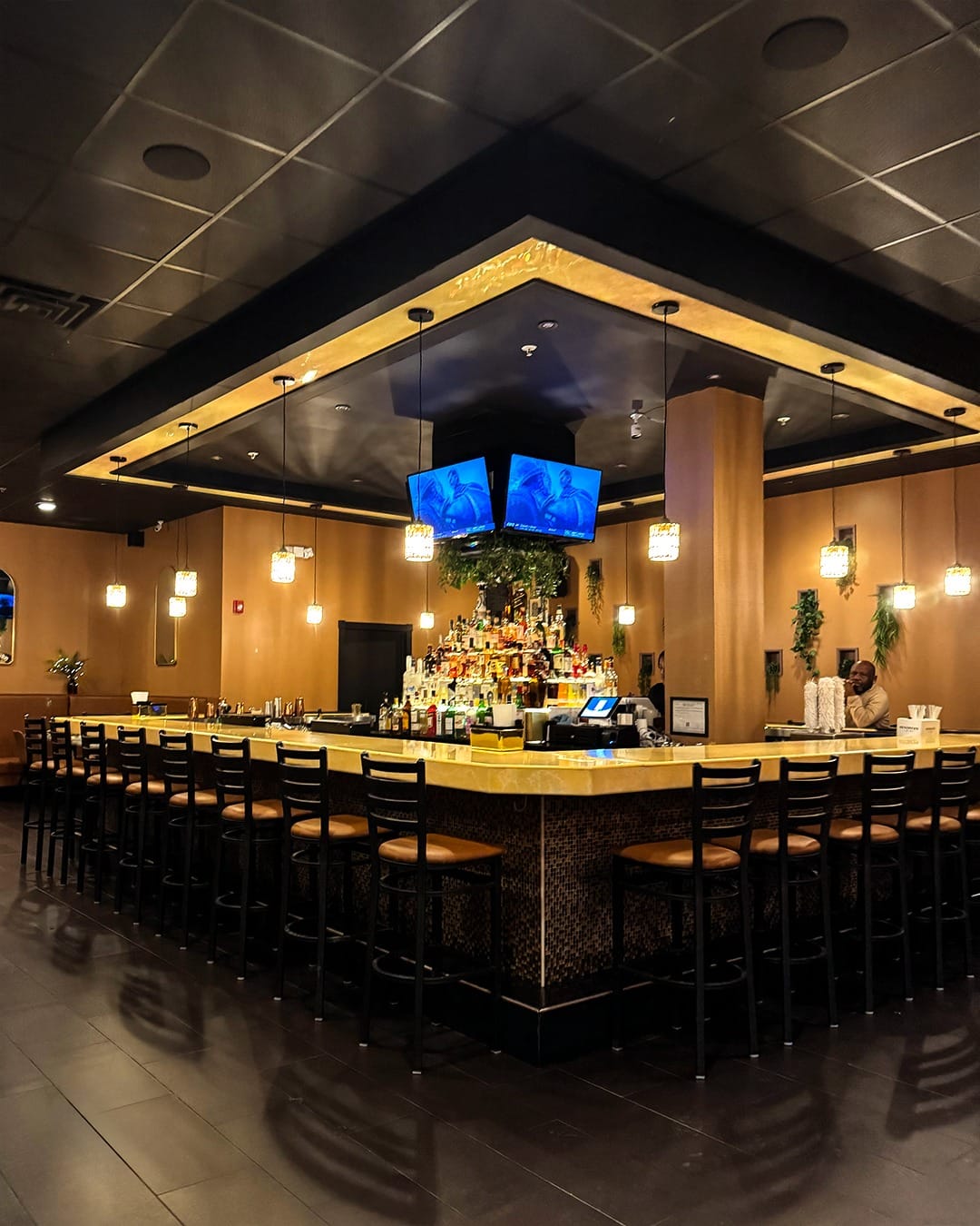 A modern bar with a square counter surrounded by high chairs, illuminated by hanging pendant lights. Bottles and TVs line the back, and a single person sits at the far end. The warm, inviting atmosphere makes Ember and Cork the perfect gathering spot.