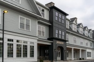 A modern gray building with white trim and large windows, featuring a central black entrance marked REGENT. The clean, well-lit exterior hints at the upscale style of Hotel Brookmere beneath overcast skies.
