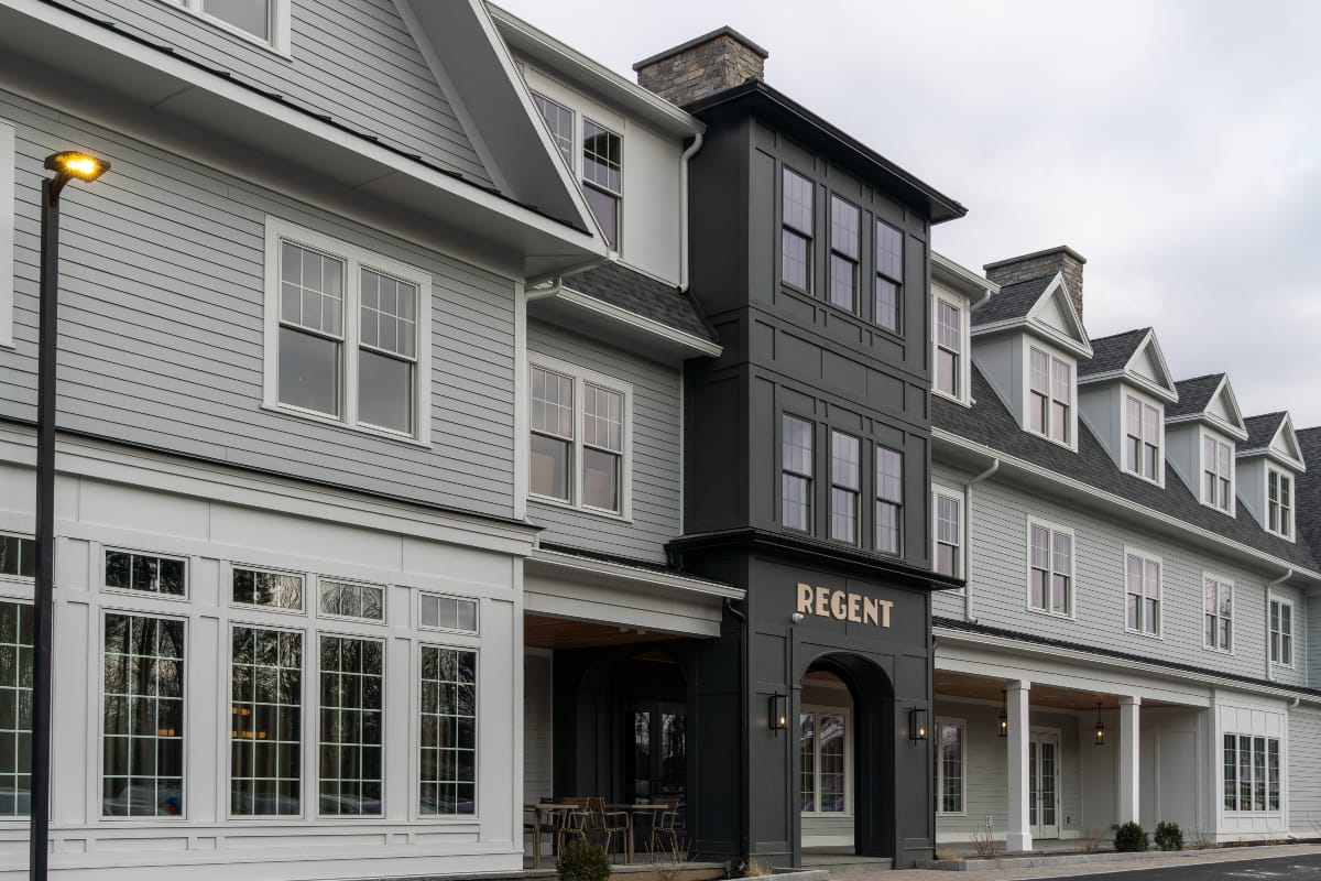 A modern gray building with white trim and large windows, featuring a central black entrance marked REGENT. The clean, well-lit exterior hints at the upscale style of Hotel Brookmere beneath overcast skies.