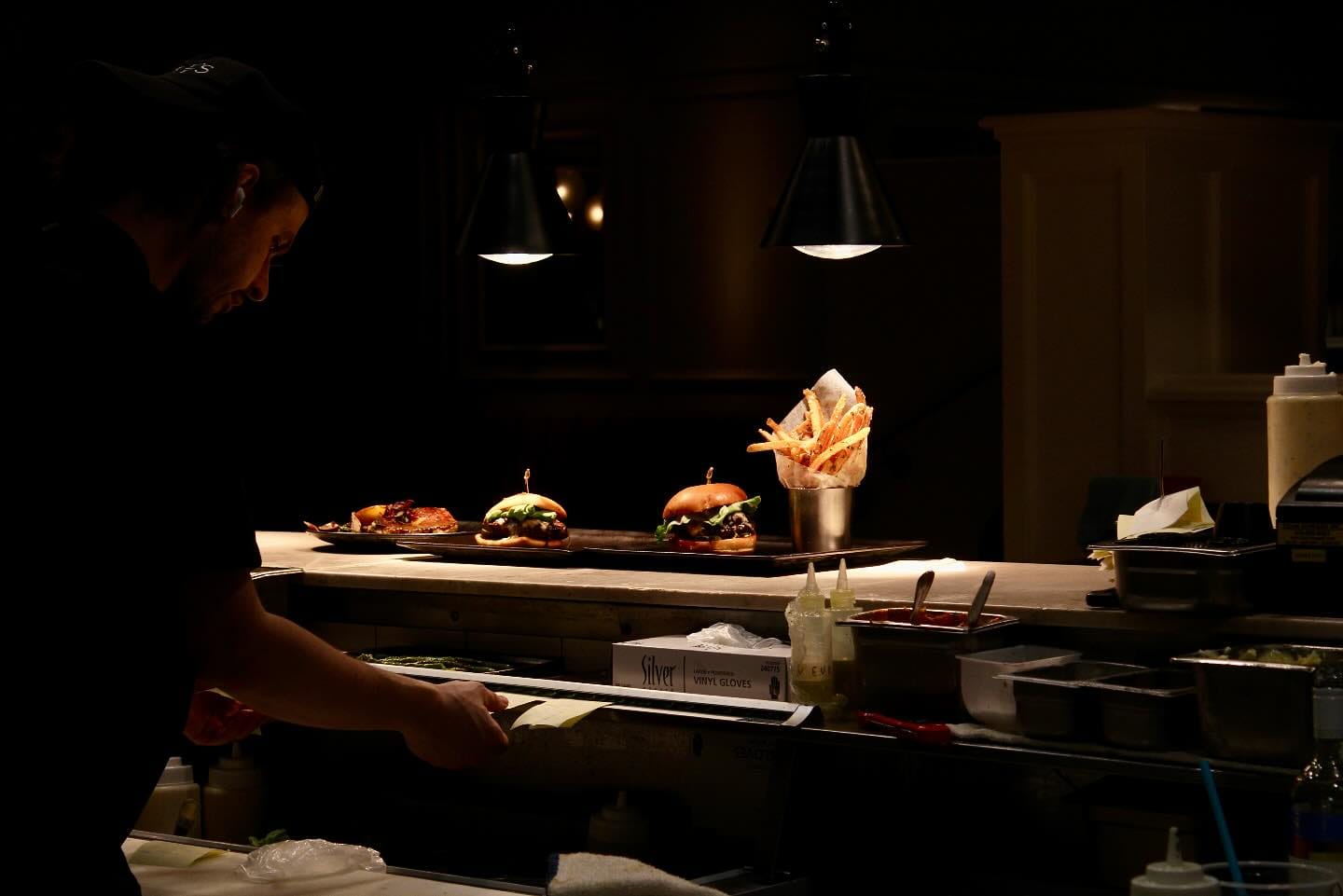 A chef works in a dimly lit kitchen at Josie's Table, preparing food. On the counter, plates with burgers and a cup holding French fries sit under a spotlight, highlighting the dishes.