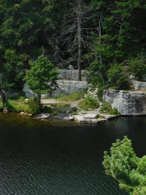 A rocky shoreline with green trees and shrubs borders a calm, dark lake. Some people are visible sitting near the water’s edge, and the scene is surrounded by dense forest.